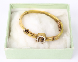 A late Victorian 15 ct yellow gold bangle featuring a lucky horseshoe set with seed pearls, hinged with a safety chain, marked '15 ct' to inner band, vendor reports in original box, total weight 10 g